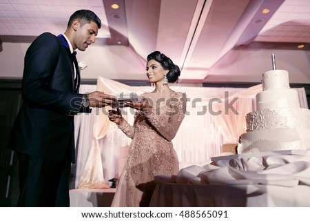 Tall groom in classy black tuxedo takes a plate with wedding cake from the hands of Indian bride