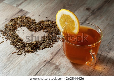 A cup of green tea standing on a wooden table. Next to a cup of heart made of dried green tea leaves