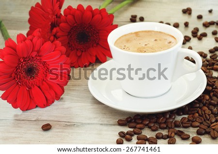 Flavored coffee in the cup and saucer is standing on a wooden table. Next to a cup of coffee, are beautiful gerbera flowers and scattered coffee beans