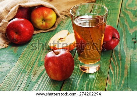 A glass of fresh juice stands on green wooden boards. Next to a glass of juice are three apples and a bag of apples that fall. bright picture