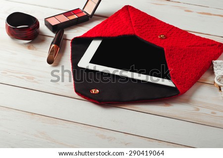 An open red lady\'s bag with tablet computer in a white wooden table.\
Near the handbags are women\'s accessories. Mascara, eye shadow, watch and bracelet
