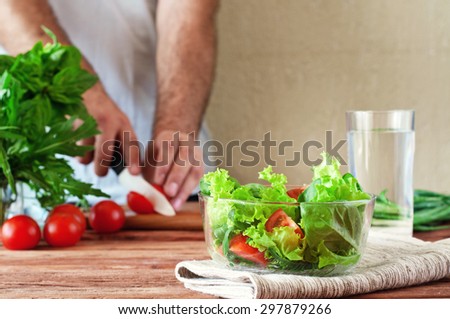 Fresh salad of summer vegetables in a deep bowl of glass. Arugula, lettuce, cherry tomatoes, basil. In the background men hand sliced cherry tomatoes on cutting board. healthy lifestyle. copy space