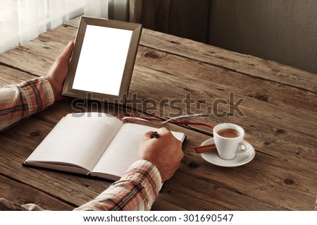 Hand of man writing something in a blank notebook. Other hand man holding an empty picture frame on wooden table. The concept of the memory of something. Top view. Copy space. Free space for text