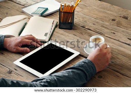 on the wooden table is a white tablet computer. Near the men hand holding a cup of black coffee. Free space for text. Copy space. Closeup. Top view