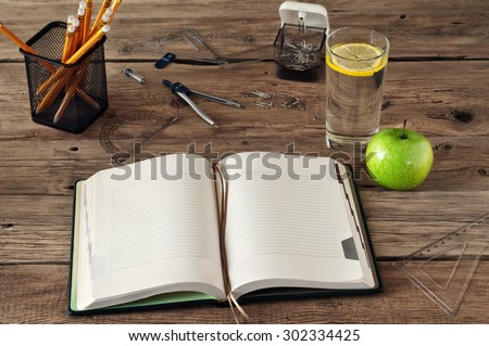 Open blank notebook on wooden background with a glass of water, apple, pencils, pens, paper clips, ruler and compass. Free space for text. Copy space. Closeup. top view