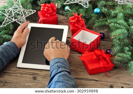 Tablet computer in men hands. Near the table with tablet computer open red gift box, christmas toys, stars and fluffy fir branches. Free space for text. top view. Copy space. Closeup