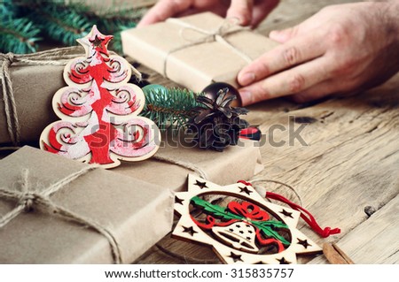 Christmas toys (wooden Christmas tree and handmade Christmas bells) with Christmas presents on a wooden table close-up. In the background, male hand putting a gift on the table