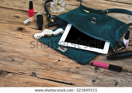 Suede womens bag with tablet computer, watch and women cosmetics (lip gloss, mascara, nail polish and spirits)  on a wooden table closeup. Top view
