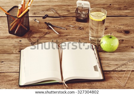 Open blank notebook on wooden background with a glass of water, apple, pencils, pens, paper clips, ruler and compass. Free space for text. Copy space. Closeup. top view. Vintage photo toning