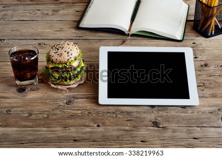 White tablet computer with a blank screen on the wooden table with a hamburger and a glass of cola with ice. Top view. Copy space. Free space for text