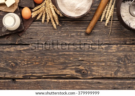 flour in a wooden bowl on dark wooden background with spikelets of wheat, eggs, milk and butter, top view with copy space. ingredients for bakery products