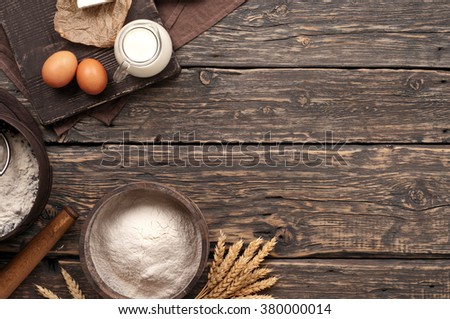 flour in a wooden bowl on dark wooden background with spikelets of wheat, eggs and milk, top view with copy space