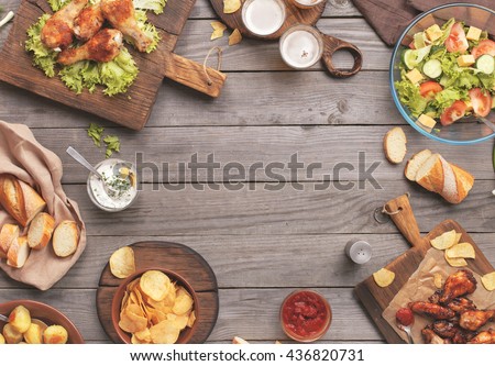 Outdoors Food Concept. On the wooden table different food with copy space, grilled chicken legs, buffalo wings, bread, salad, potatoes, potato chips and beer