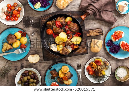On the wooden table a different food, fried meat with vegetables in a pan, salad and snacks, top view