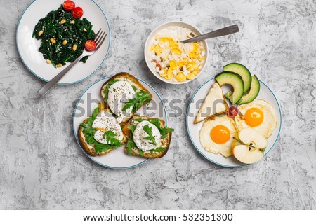 Delicious breakfast - fried egg, poached eggs, avocado, spinach salad, muesli and cheese sandwiches on light surface, top view