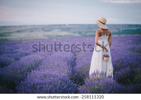 Beautiful young woman in wicker hat and white dress posing in a lavender field with small wicker basket in her hand