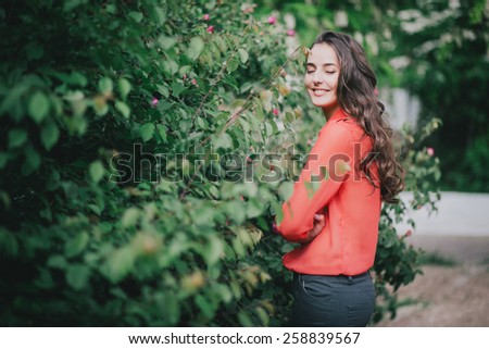 Beautiful girl with long curly hair in a red chiffon shirt posing in a green rose garden