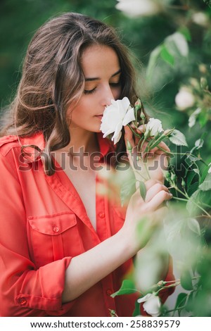 Beautiful girl with long curly hair in a red chiffon shirt smelling a rose in a green rose garden
