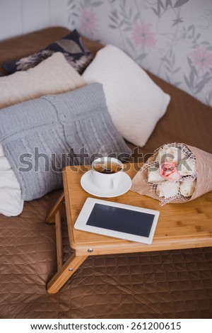Wooden tray with tea, flowers and e-book standing on a bed with decorative pillows in a bedroom. Cozy scandinavian interior