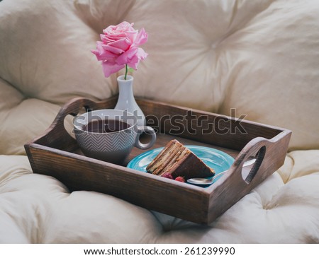 Cake with fresh strawberries and hot tea and a rose in a small vase standing on a wooden tray on a white couch. Still life
