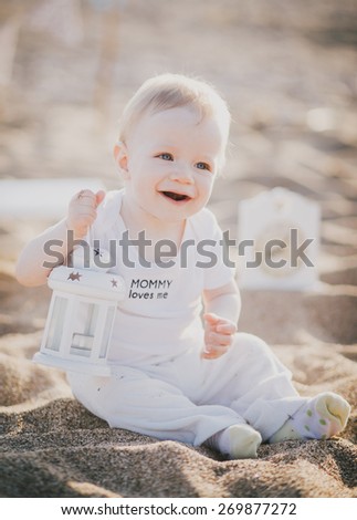 Little baby-boy in a cute suit sitting on a sand at the beach with white lantern. Cute blue flags on the background