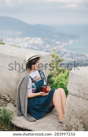 Young fashion woman sitting on a top of the mountain drinking tea with beautiful sea and hills view on the background