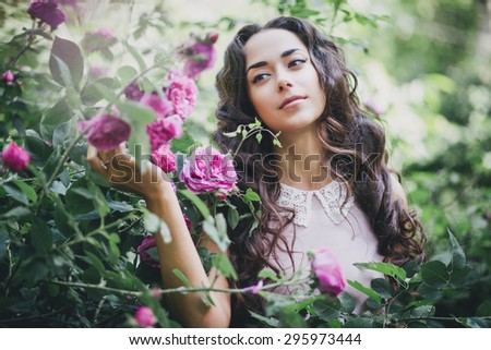 Beautiful young woman in a pink dress with long curly hair posing near roses in a garden