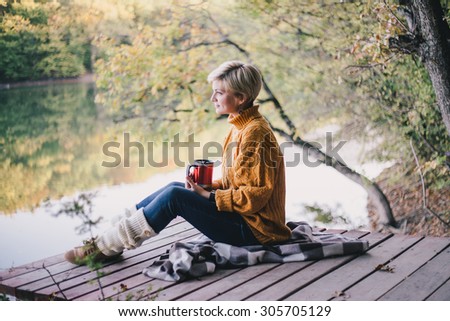 Young beautiful blond dressed in oversize knitted sweater and jeans sitting near lake with cup of hot tea
