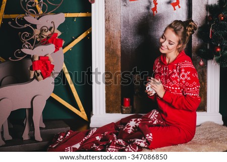 Young beautiful woman in a red warm pajamas with scandinavian ornaments sitting near decorative fireplace and drinking hot tea or cocoa. Christmas mood