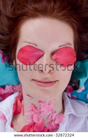 Portrait of young woman with pink petals in front of eyes. She is looking through flowers and smiling. Some petals laying on her neck. White blouse. Shallow depth of field.