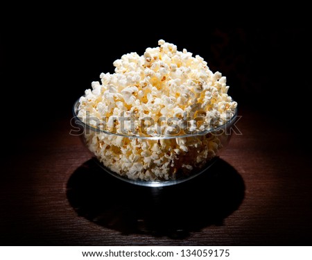 Many crunchy popcorn in glass bowl and black shadow around on table, black background. Traditional food prepared and ready to eat, standing on dark table in light flux, horizontal orientation.