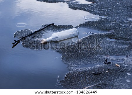 One empty glass bottle garbage on melting ice on lake surface late winter season, raining weather in Poland. Winter detritus at frozen water, blue toned. Horizontal orientation, nobody.