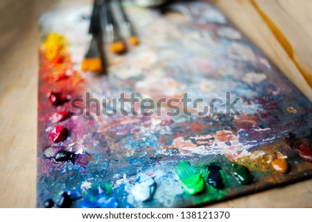 Many oil mixed paints on wooden palette with brushes blurred behind lying on wooden board, horizontal orientation, nobody.