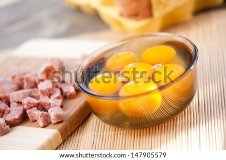 Six raw egg yolks in glass bowl and chop sausage lying on board, preparing scramble eggs, horizontal orientation, nobody, objects in studio shot.