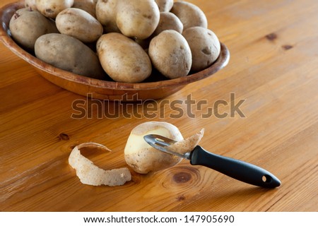 Potato peeler and peelings of tuber, many vegetables lying on bamboo bowl on wooden table, horizontal orientation, nobody, daylight from window.