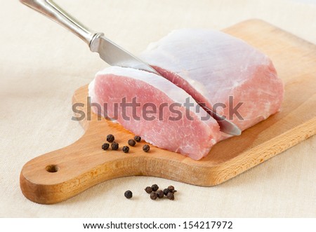 Knife in pork meat for cutlets or schabowy kotlet, chopping fresh portion of meat and grains of pepper lying on trencher board, ready to prepare cuisine. Nobody, horizontal orientation.