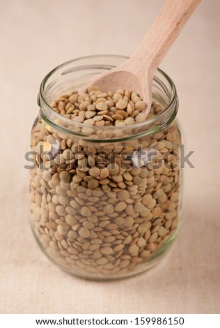 Green lentils seeds and wooden spoon in jar of glass, plenty healthy raw grains in studio shot, vertical orientation, nobody.