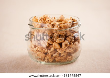 Crisp popped wheat grains in glass jar on beige material, healthy puffed cereal food staple in studio shot, horizontal orientation, nobody.