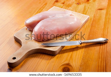 Single raw whole chicken breast and knife lying on wooden board and table ready to cut, uncooked meat in horizontal orientation, nobody, studio shot.
