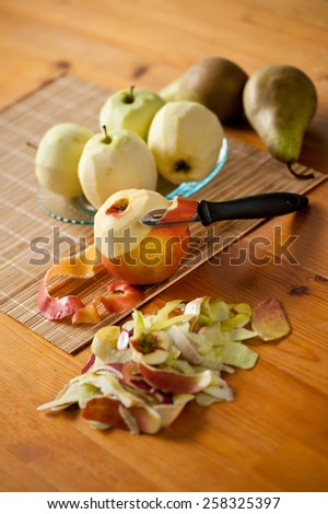 Peel ripe fruits apples and pears on glass plate on bamboo mat, peelings and whole fruits lying on wooden table, nobody, vertical orientation.