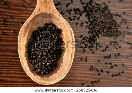 Black Nigella Sativa dry seeds portion on wooden spoon and spilled around food macro, Black cumin raw spice heap in day light, horizontal orientation, nobody. Polish name czarnuszka siewna.