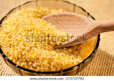 Bulgur wheat groats portion in bowl, dried burghul cereal food portion and wooden spoon in glass bowl lying on beige mat, grains healthy food heap in hard day light, horizontal orientation, nobody.