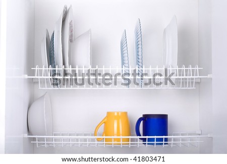Open white dish draining closet with wet dishes of glass and ceramic, plates, bowls, covers and mugs objects drying inside on rack made of plastic coated steel wire and open bottom