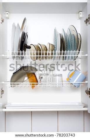 Open white dish draining closet with wet dishes of glass and ceramic, plates, bowls, covers and mugs fragile objects drying inside on rack made of plastic coated steel wire and open bottom