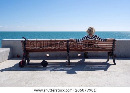 Young woman sitting on the bench near sea with black scooter/push stand-up scooter/kick scooter.
