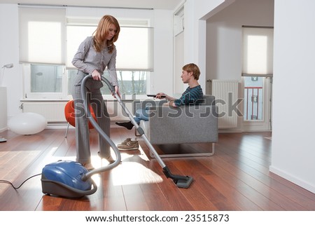 Business woman is vacuum cleaning while the lazy boyfriend is sitting on the couch watching tv