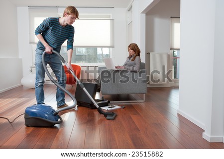 Man is vacuum cleaning while his girlfriend is working on the couch