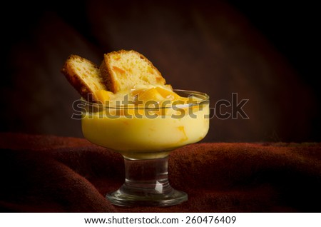 a new style trifle placed in a glass dish stacking together 2 thin slices of orange cake, some mango cubes and an orange custard, shot in studio on a warm reddish brown background