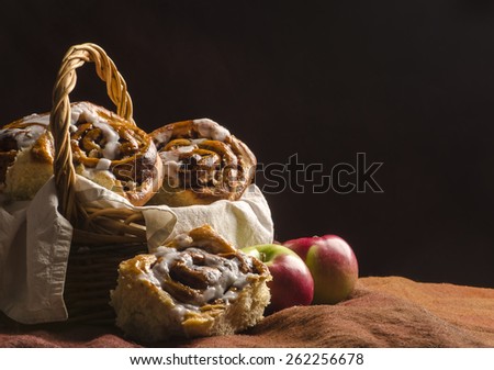 a wicker basket layered with an off-white cloth is filled with some chelsea apple brioche with one brioche place on the side of the basket with two apple. Shot on a warm dark backdrop