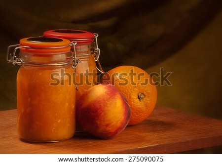 2 glass jars filled with orange-peach marmalade with a fresh orange and a fresh peach placed on the side, shot in studio on a dark brown backdrop